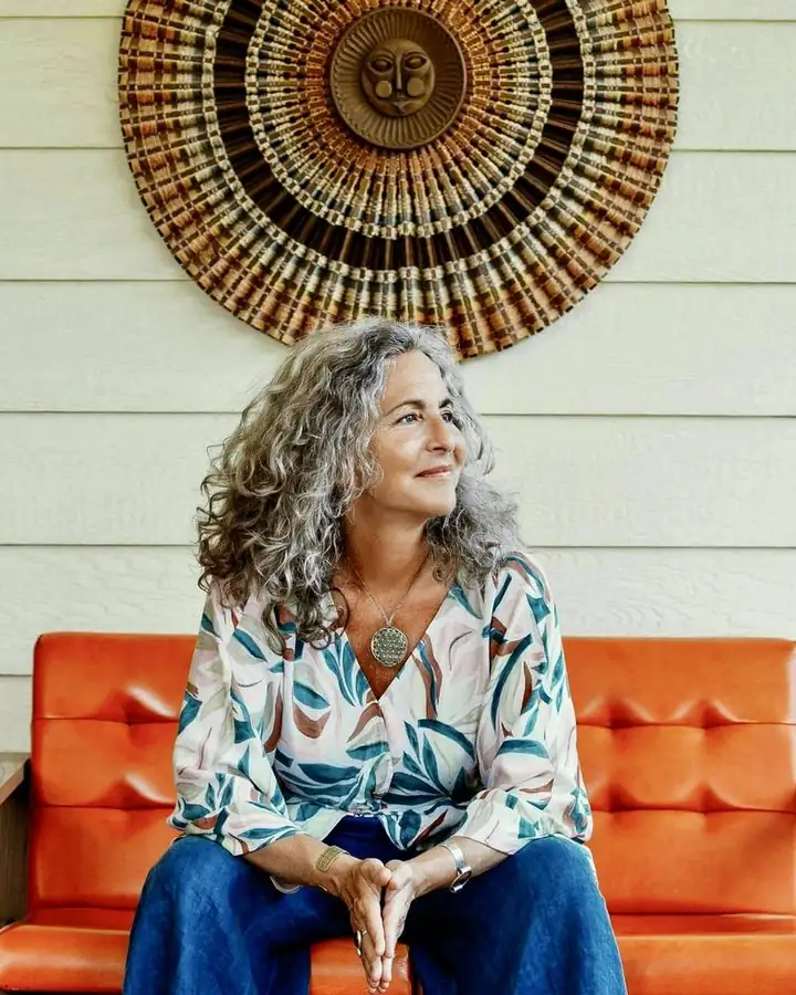 Heidi Feinstein seated on an orange sofa beneath a woven sun, smiling and looking to the side